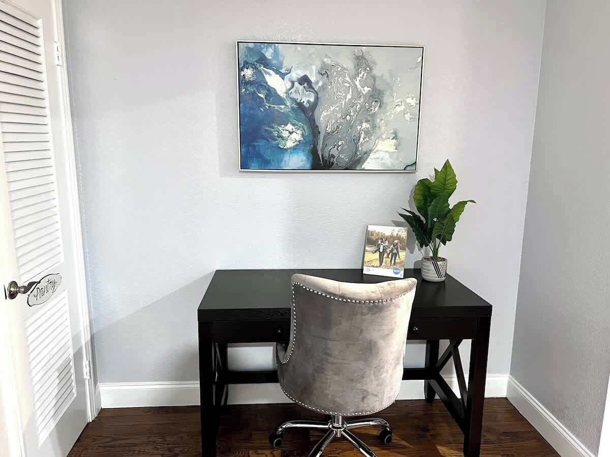 A room with a dark wooden desk, a grey upholstered chair, a framed picture on the wall, and a potted plant on the desk.