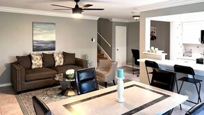 Ein modernes Wohnzimmer mit Sofa, Couchtisch und Sesseln. Es gibt einen Esstisch mit Kerze. Ein Deckenventilator ist sichtbar, und im Hintergrund befindet sich ein Küchenbereich.