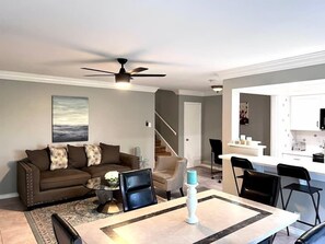 A modern living room with a sofa, coffee table, and chairs. There is a dining table with a candle. A ceiling fan is visible, and a kitchen area is in the background. - Modern 2BD Pool  Six Flags Grand Prairie GP08B (Grand Prairie)