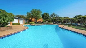 Ein klarer, blauer Swimmingpool mit gefliestem Rand, umgeben von einer angelegten Grünfläche mit Bäumen und einem Gebäude im Hintergrund.