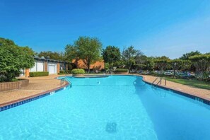 A clear blue swimming pool with a tiled edge, surrounded by a landscaped area with trees and a building in the background. - Modern 2BD Pool  Six Flags Grand Prairie GP08B (Grand Prairie)