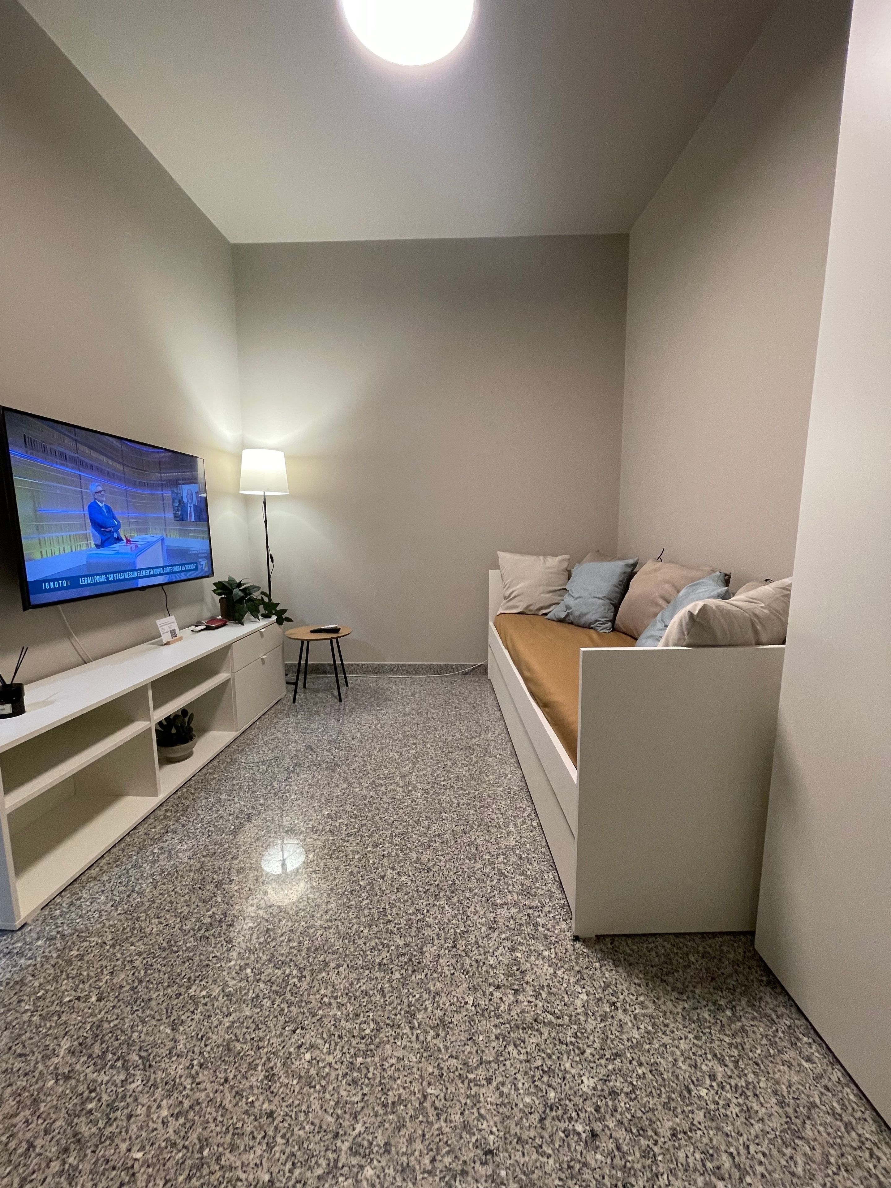 Ein kleiner, moderner Wohnraum mit einem Wand-Fernseher, einem weißen Sofa mit Kissen, einem kleinen Beistelltisch und einer Lampe.