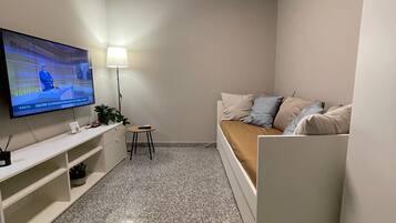 Ein kleiner, moderner Wohnraum mit einem Wand-Fernseher, einem weißen Sofa mit Kissen, einem kleinen Beistelltisch und einer Lampe.