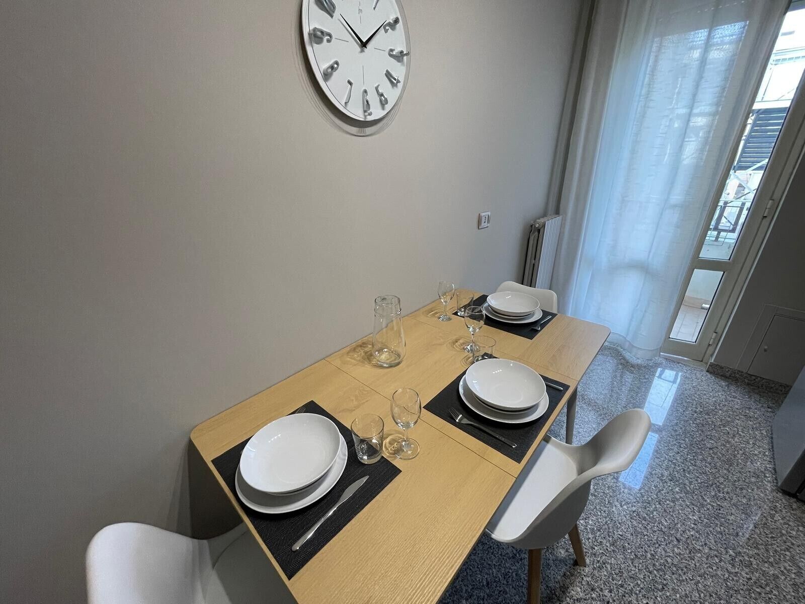 Un comedor con una mesa de madera dispuesta para dos personas, un reloj de pared y vistas al exterior a través de una ventana.