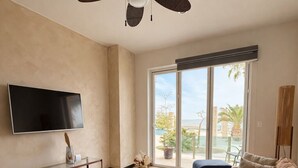 Una sala de estar moderna con ventilador de techo, televisor de pantalla plana, mesa de centro de vidrio, sofá y vistas al exterior a través de una puerta corrediza de vidrio.