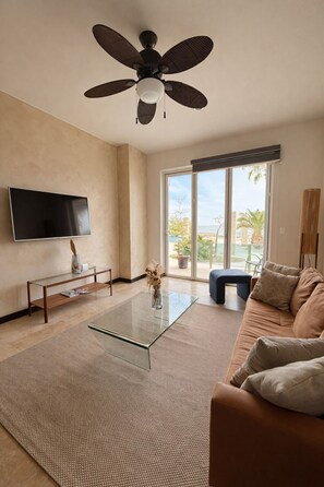 Ein modernes Wohnzimmer mit Deckenventilator, Flachbildfernseher, Glastisch, Sofa und Blick ins Freie durch eine Glastür.