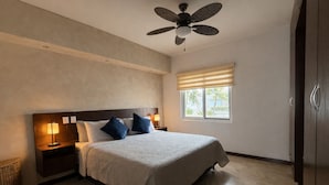 Un dormitorio con cama, mesitas de noche, ventilador de techo, ventana con persianas y vista a palmeras.