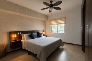 Ein Schlafzimmer mit einem Bett, Nachttischen, einem Deckenventilator, einem Fenster mit Jalousien und Blick auf Palmen.