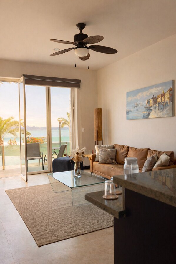 A modern living room with a glass coffee table, a brown leather sofa, and a ceiling fan. - Oceanfront Suite (El Centenario)