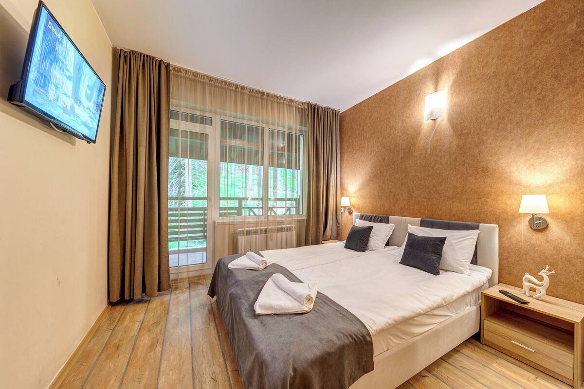 A hotel room with a large bed, a TV mounted on the wall, a window with curtains, and a nightstand with a lamp and a decorative item.