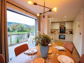 A dining area with a table set for two, a vase of flowers, and a view of a river and greenery outside. - Mountain View Cottage (Llanrwst)