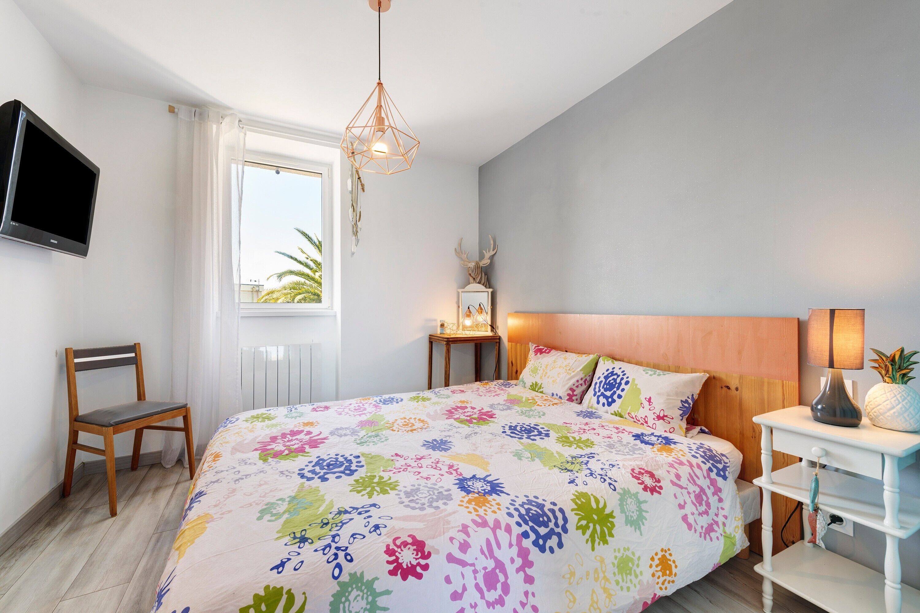 A bedroom with a bed, a chair, a television, a window, and a bedside table with a lamp.