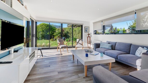 Un salon moderne doté d’un téléviseur à écran plat, d’un canapé gris, d’une table basse blanche et de grandes fenêtres offrant une vue sur la verdure.