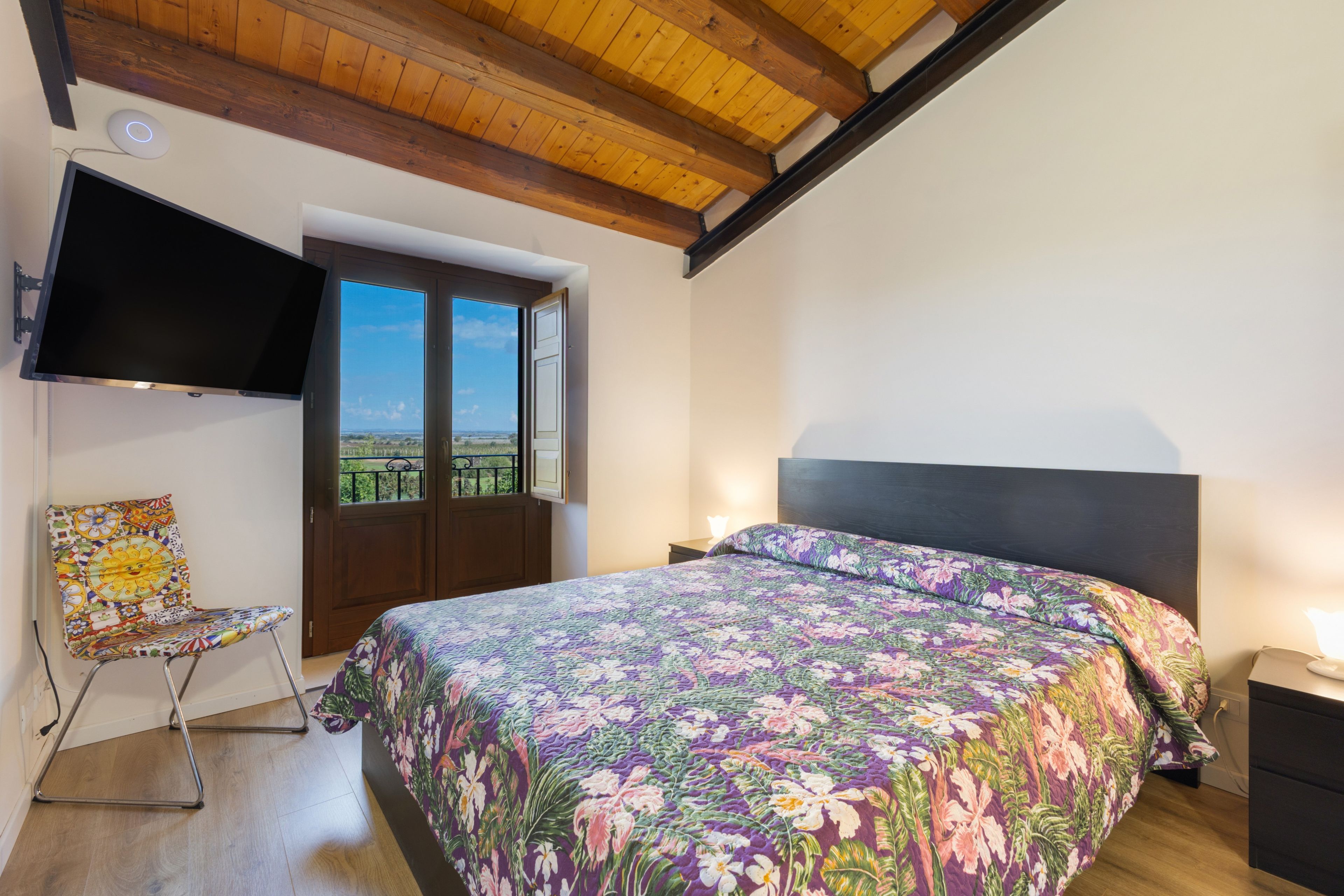 A bedroom with a bed, a TV mounted on the wall, a chair, and a window with a view.