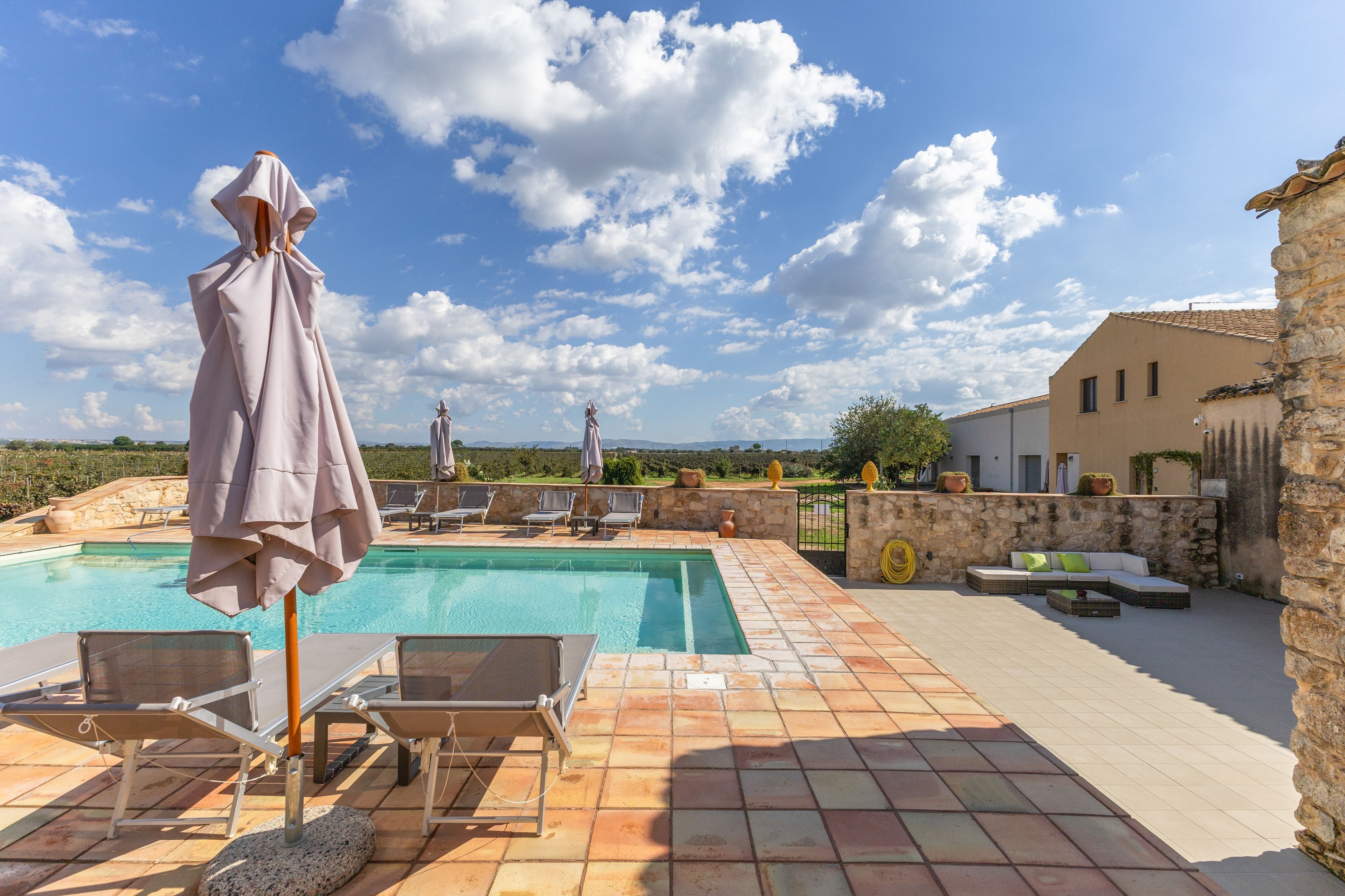 A swimming pool with lounge chairs and umbrellas, a stone wall, and a view of the countryside.