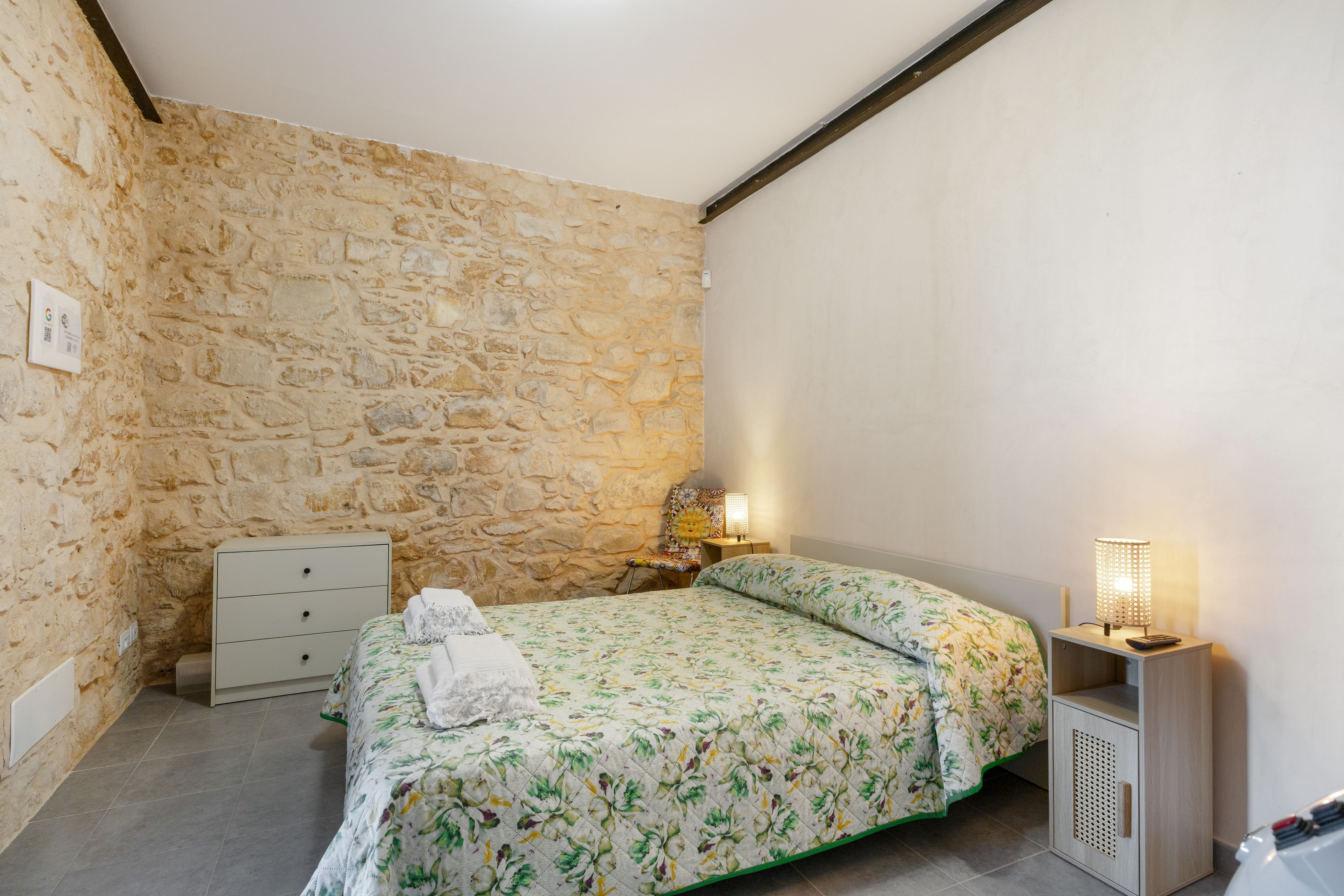 Ein Schlafzimmer mit Steinwand, einem Bett mit floralem Überwurf, einem Nachttisch mit Lampe und einer Kommode.