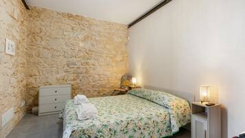 Ein Schlafzimmer mit Steinwand, einem Bett mit floralem Überwurf, einem Nachttisch mit Lampe und einer Kommode.