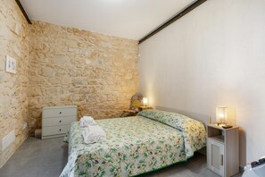 Ein Schlafzimmer mit Steinwand, einem Bett mit floralem Überwurf, einem Nachttisch mit Lampe und einer Kommode.