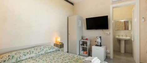 Et hotelværelse med seng, fjernsyn, skrivebord med lampe, lille køleskab, kaffemaskine og badeværelse med vask og spejl.