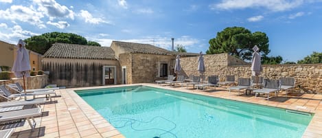 A swimming pool with lounge chairs and umbrellas, surrounded by stone buildings and trees.
