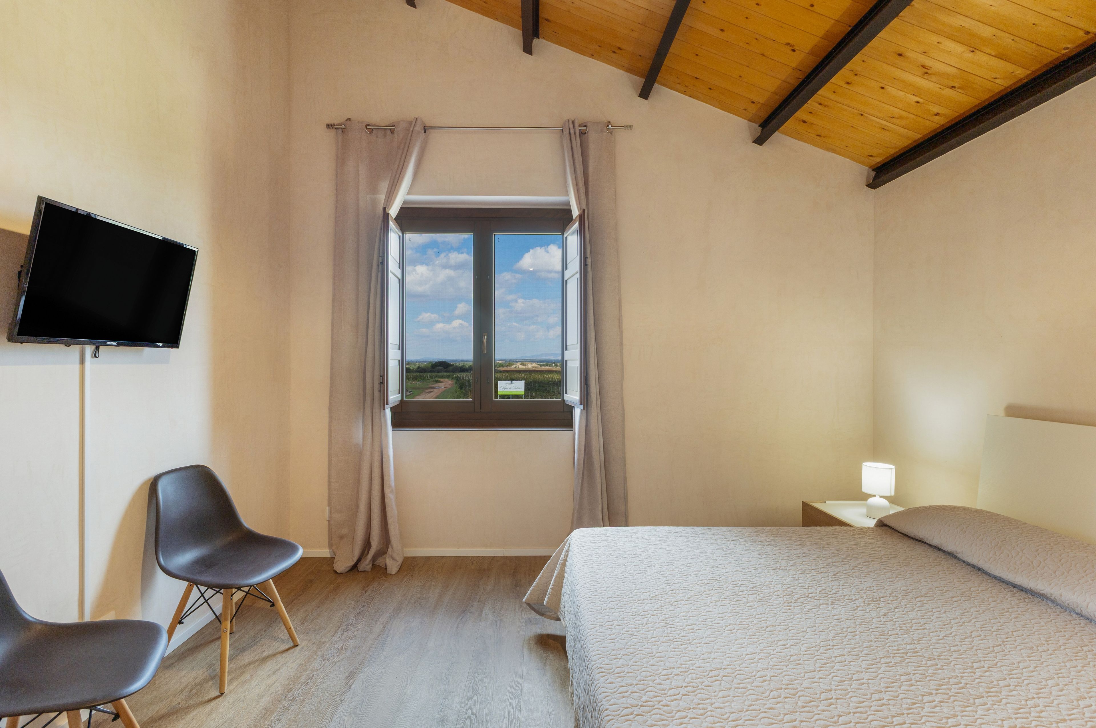 Un dormitorio con cama, una silla, un televisor, un ventanal con cortinas y una mesita de noche con lámpara.