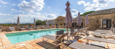 A poolside area with lounge chairs and umbrellas, a swimming pool, and a stone building in the background.