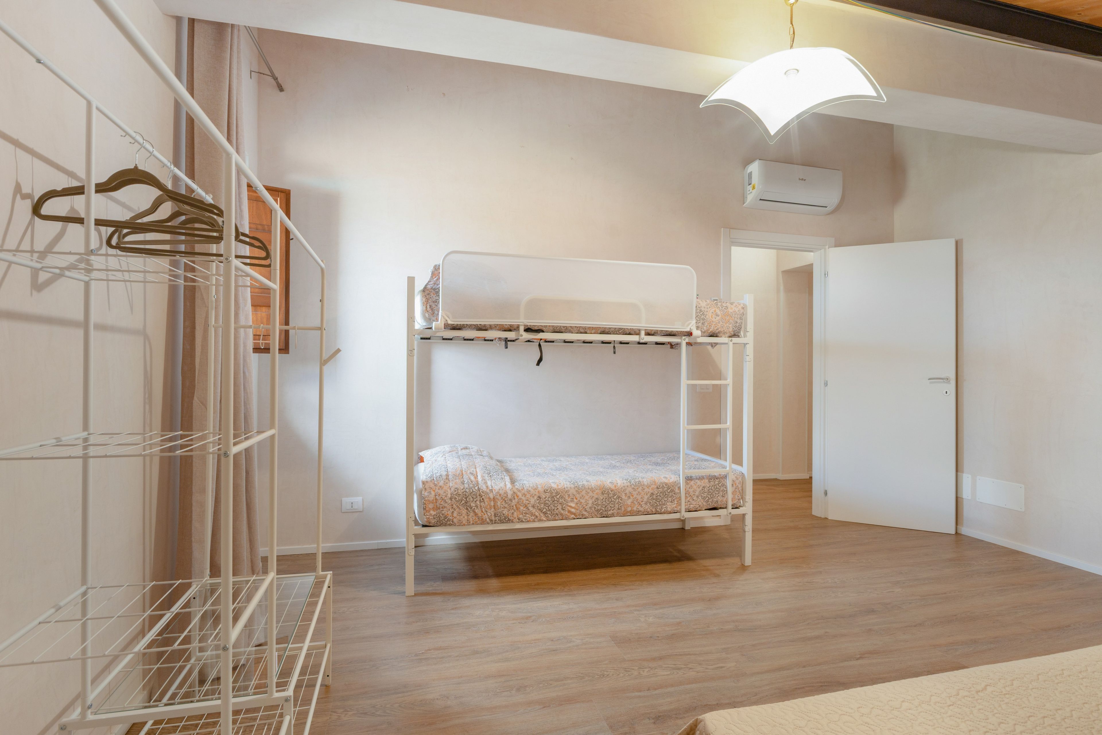 Ein Etagenbett mit weißem Kopfteil, ein Metallregal mit Kleidung und eine Tür im Hintergrund.