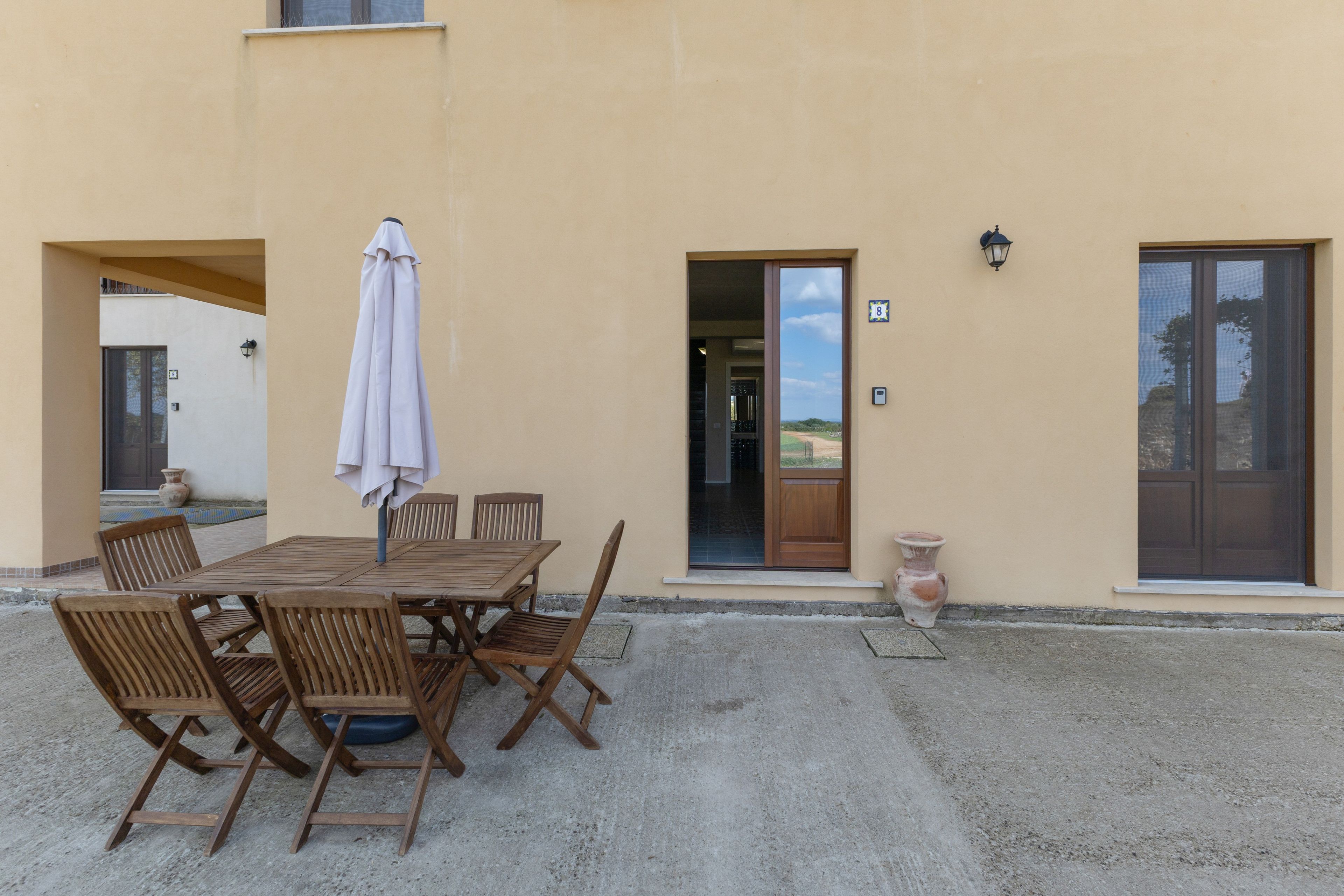 Eine Terrasse im Freien mit Holztisch und -stühlen, einem weißen Sonnenschirm und Blick auf die Landschaft durch die offene Tür.