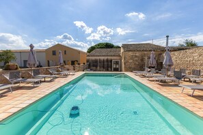 Ein rechteckiger Pool mit klarem Wasser, umgeben von Liegestühlen und Sonnenschirmen, mit Gebäuden und einer Steinmauer im Hintergrund.