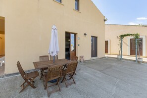 Un patio con una mesa y sillas de madera, un paraguas blanco y un edificio de fachada color beige.