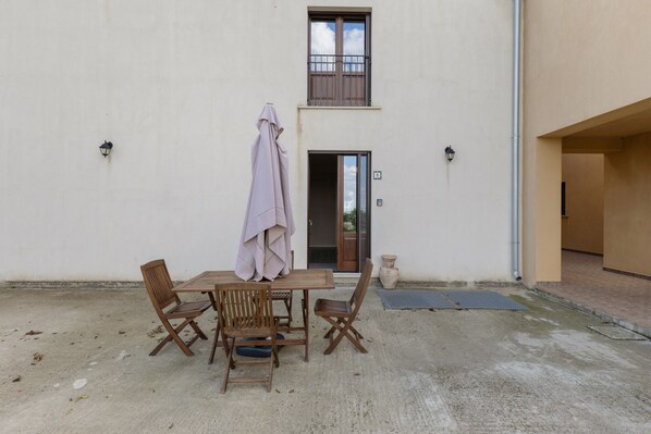 Un patio exterior con una mesa y sillas de madera, un paraguas cubierto y la entrada de un edificio con una puerta y ventanas.