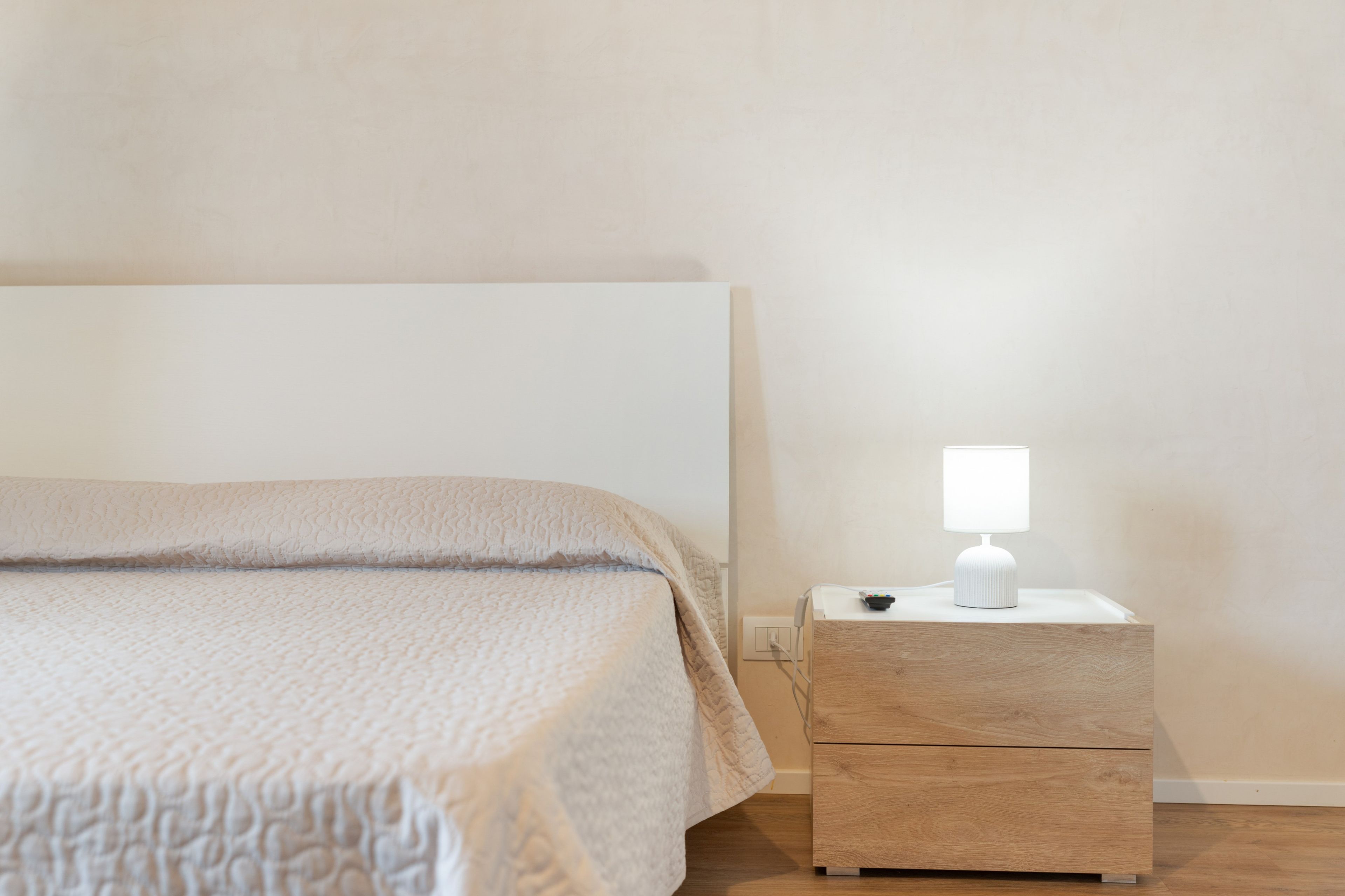 Een bed met een geruite deken, een nachtkastje met een lamp en een stopcontact.