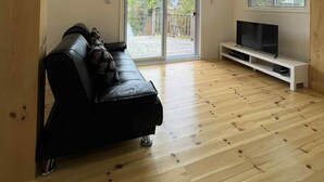 Ein geräumiges Wohnzimmer mit einer schwarzen Ledersofa, einem Flachbildfernseher auf einem weißen Standfuß, Holzböden und großen Schiebeglasfenstern zu einem Außenbereich.