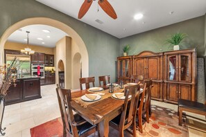 Un comedor con una mesa de madera dispuesta para seis personas, rodeada de sillas. Se observa una cocina al fondo con electrodomésticos modernos y un arco que conduce a otra habitación.
