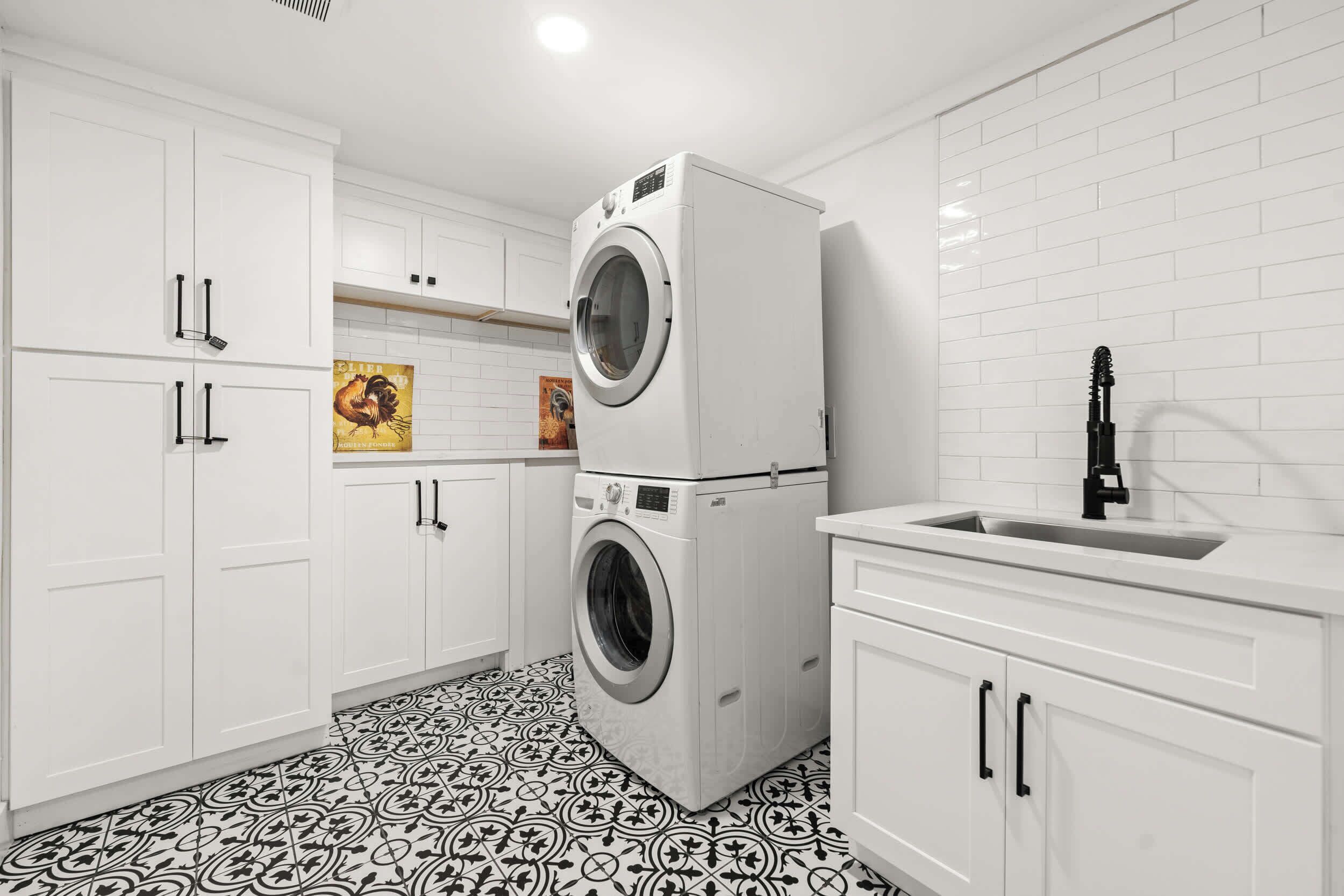 A laundry room with a stacked washer and dryer, white cabinets, a sink, and patterned floor tiles.