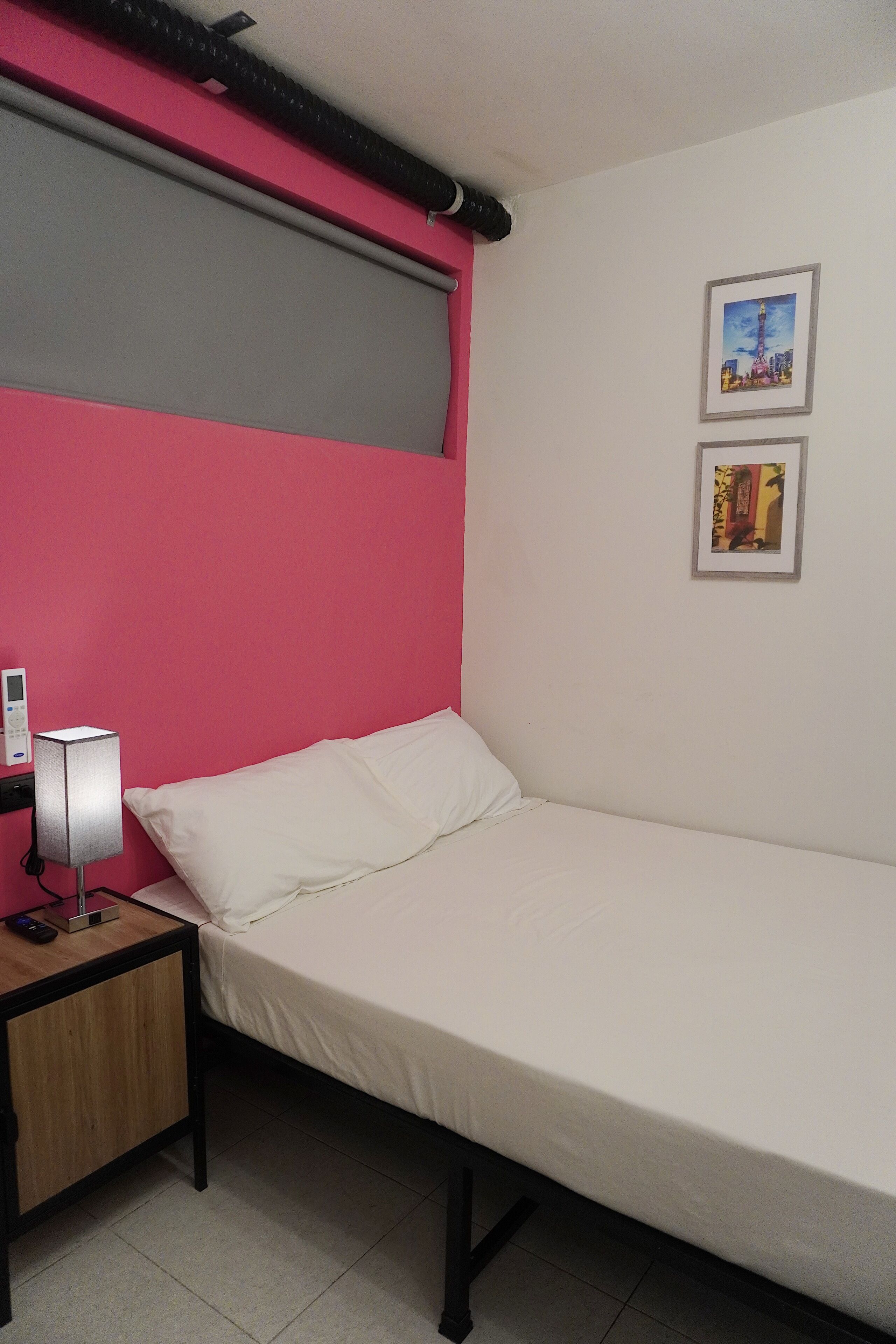 A single bed with a white mattress and pillows, a bedside table with a lamp, and a wall-mounted air conditioning unit.
