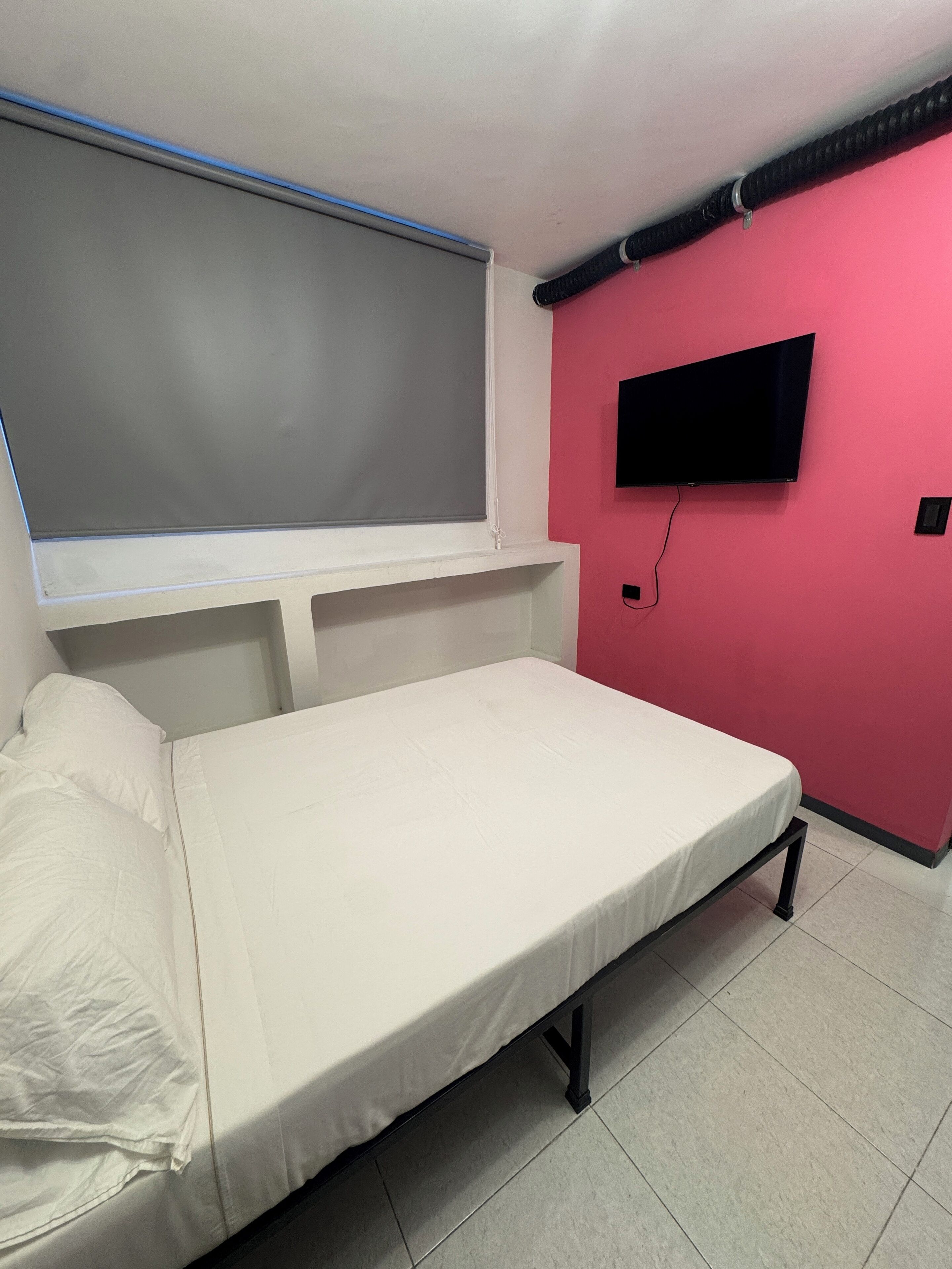 Una habitación pequeña con cama individual, ventana, televisor de pantalla plana montado en la pared y suelo de baldosas.