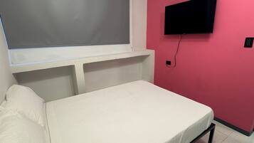 Una habitación pequeña con cama individual, ventana, televisor de pantalla plana montado en la pared y suelo de baldosas.