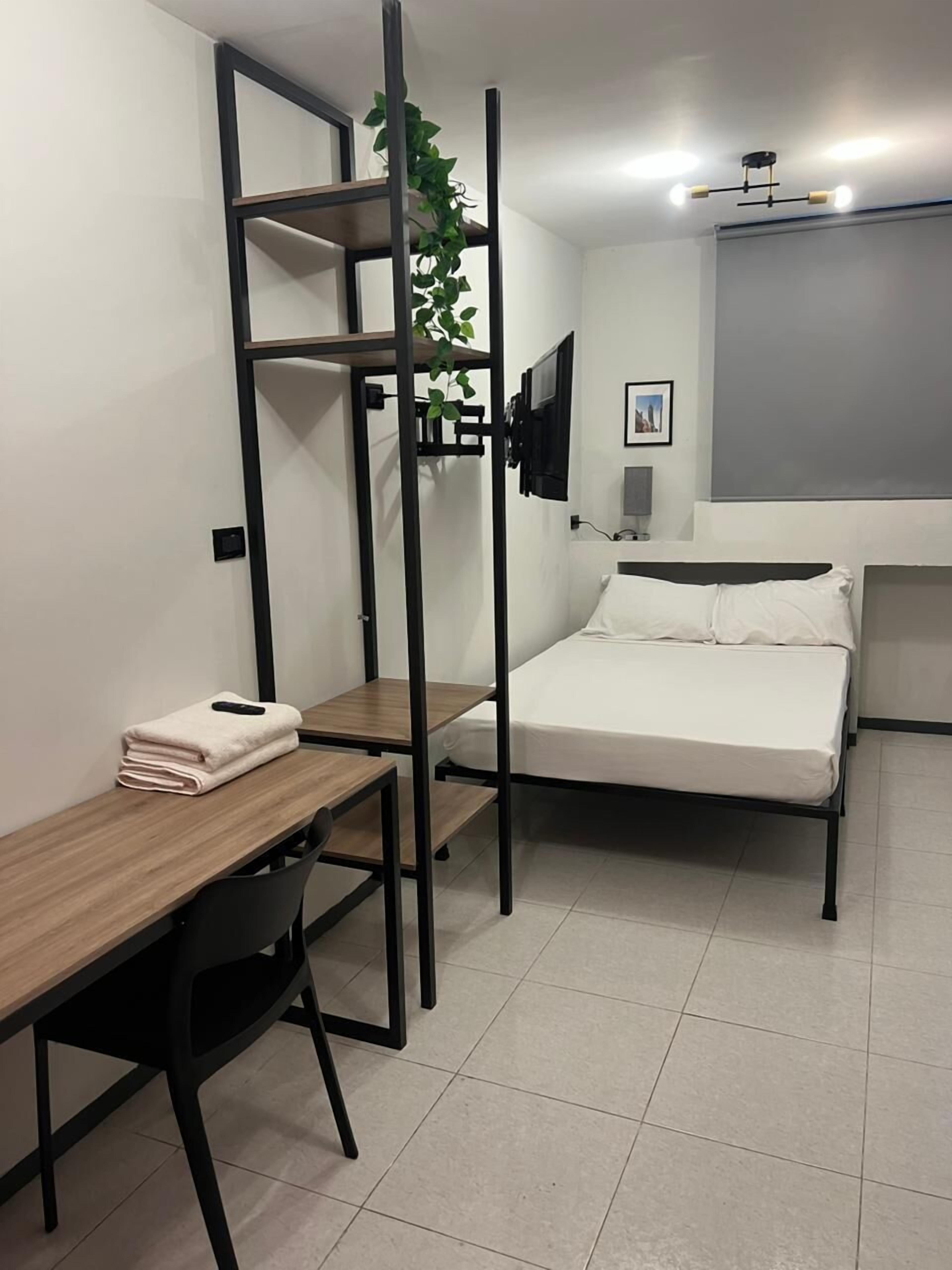 Habitació petita amb llit, escriptori, cadira, televisió penjada a la paret i una planta sobre una estant.