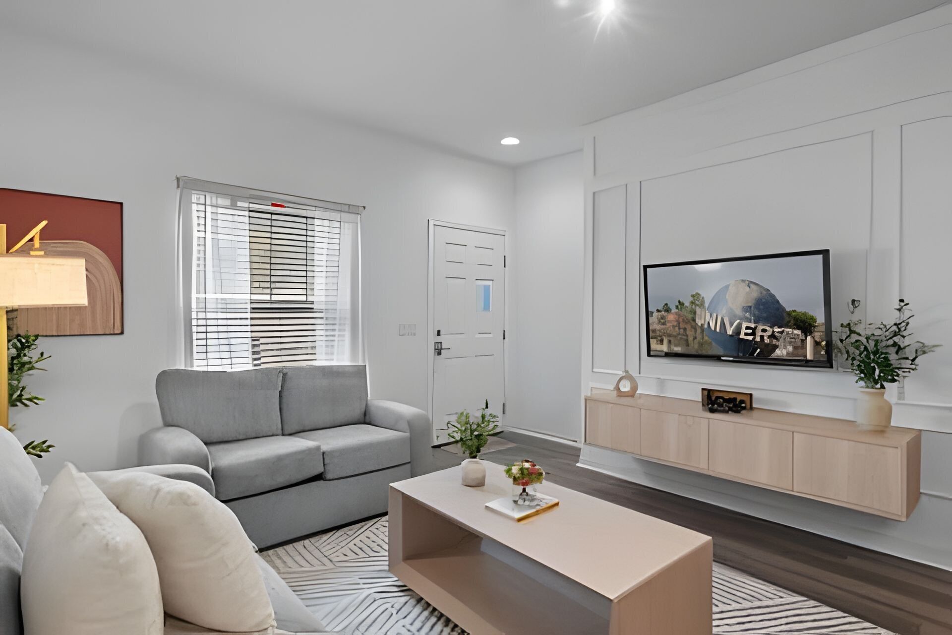 Ein modernes Wohnzimmer mit einem grauen Sofa, einem Couchtisch, einem flachbildfernseher an der Wand und einem Fenster mit Jalousien.