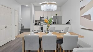 Un'area pranzo moderna con un tavolo in legno e quattro sedie grigie, un lampadario e una cucina sullo sfondo.