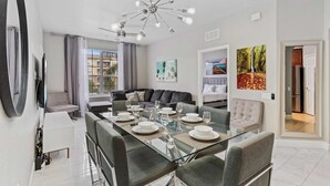 A modern dining area with a glass table set for six, a sofa, and a ceiling light fixture. - Spacious 3BR Condo at Vista Cay Resort (Orlando)