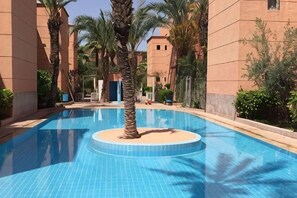 Une piscine dotée d’un îlot circulaire, de palmiers et d’un bâtiment à la façade beige.