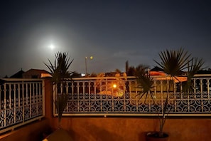 A gated balcony with a metal fence, potted plants, and a view of a building with lights in the background. - Riad with Swimming Pools (Marrakech)