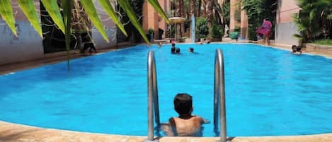 An indoor swimming pool with clear blue water, surrounded by palm trees and a tiled floor.