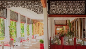 Uma área de estar coberta ao ar livre, com mesas, cadeiras, plantas em vasos e vista para o exterior.