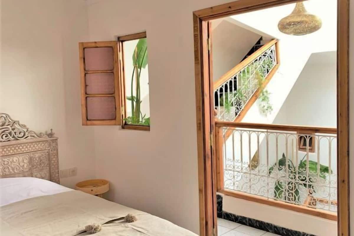 Un dormitorio con cama, mesita de noche, espejo y vista a un balcón con barandilla.