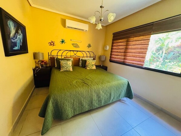 Une chambre avec une couvre-lit verte, une table de chevet avec une lampe, un climatiseur fixé au mur et une fenêtre avec des stores.