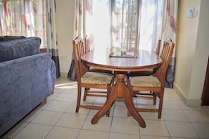 A dining area with a round wooden table and four chairs, a blue sofa, and curtains with floral patterns. - Stay happy at Raz Homes (Diani Beach)