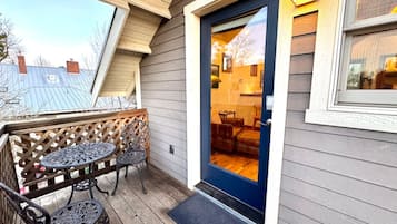 Une terrasse en bois avec deux tables et des chaises en métal, une porte bleue donnant sur un espace intérieur, et une vue sur les maisons voisines.